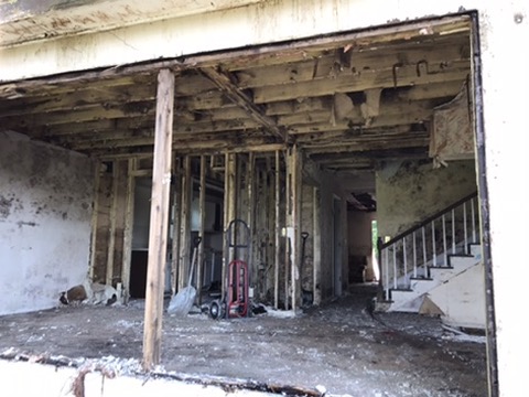 Demolition seen through an opening in the exterior wall: blackened studs and joists around a staircase, subfloor stripped.