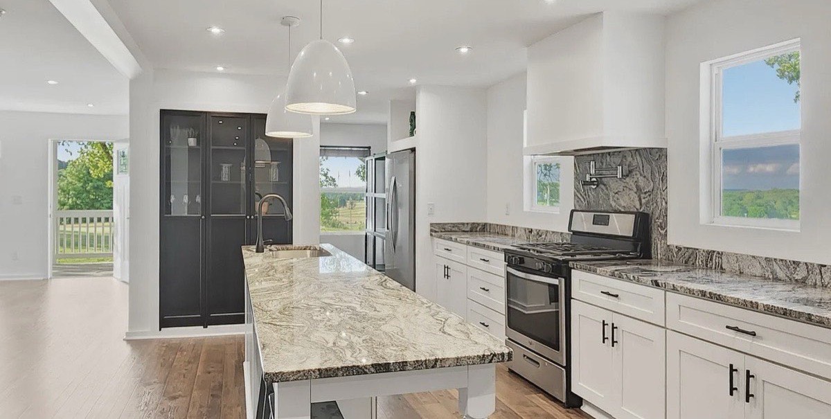 The finished kitchen: a long stone-topped island, white cabinetry, gas range and stone backsplash, with the great room visible through a wide opening at the left.