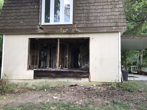 A rectangular opening cut through the first-floor exterior wall, siding and sheathing removed, rotted band joist exposed at the sill.